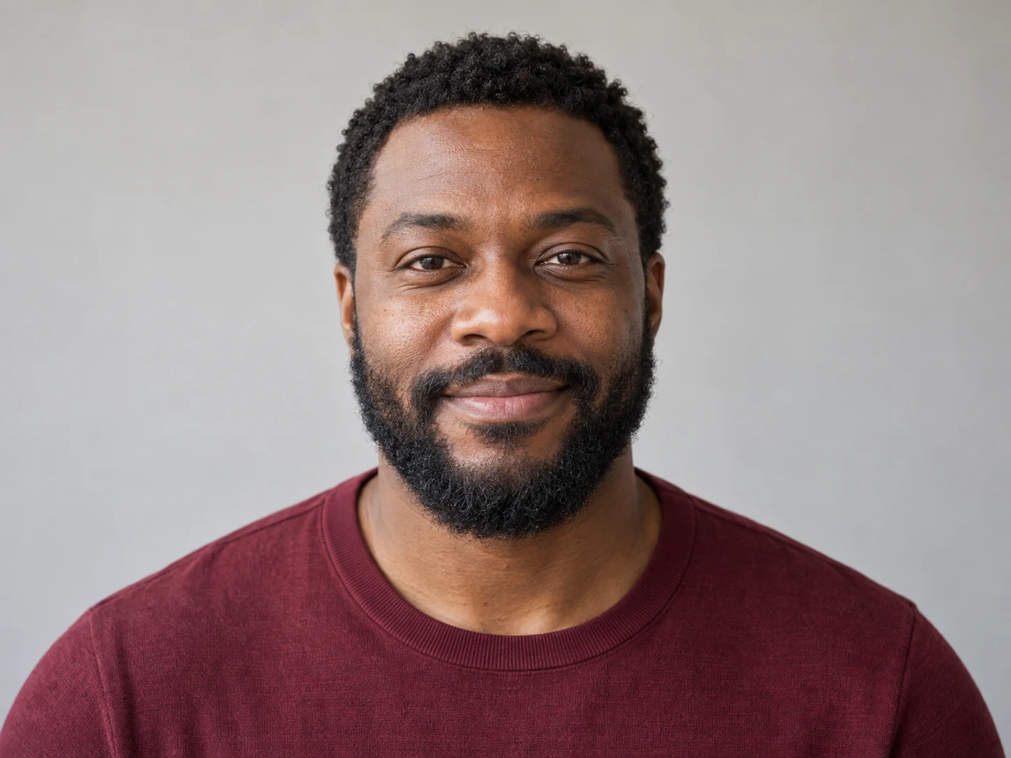 Adult Black man with a dense short beard and moustache, a burgundy shirt, and soft window light.